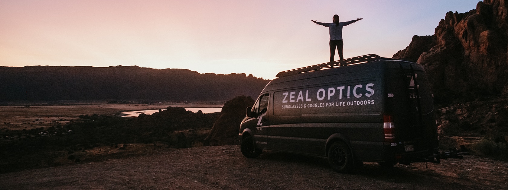 a person standing on top of a Zeal Optics van near a rocky landscape
