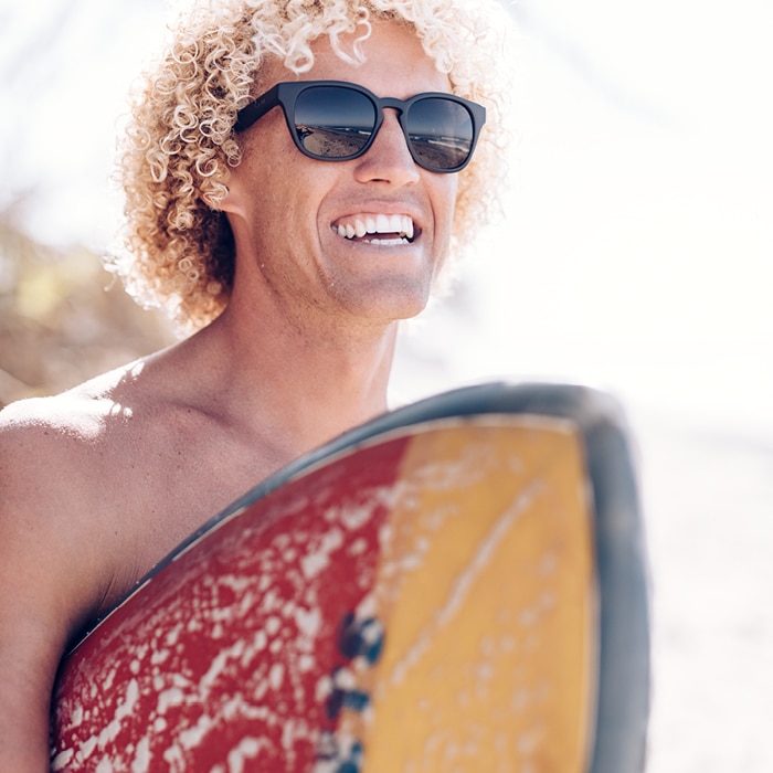 a smiling man wearing Zeal sunglasses and holding a surfboard