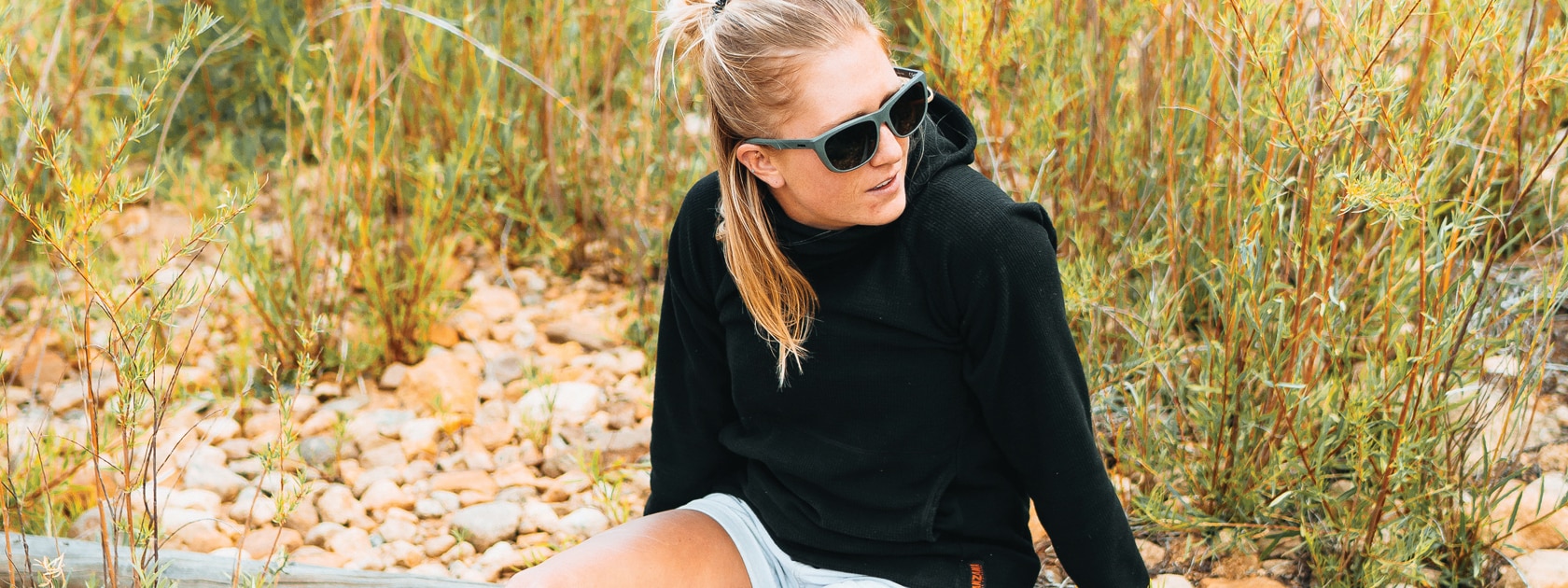 woman in a black hoodie and sunglasses sits near tall grass