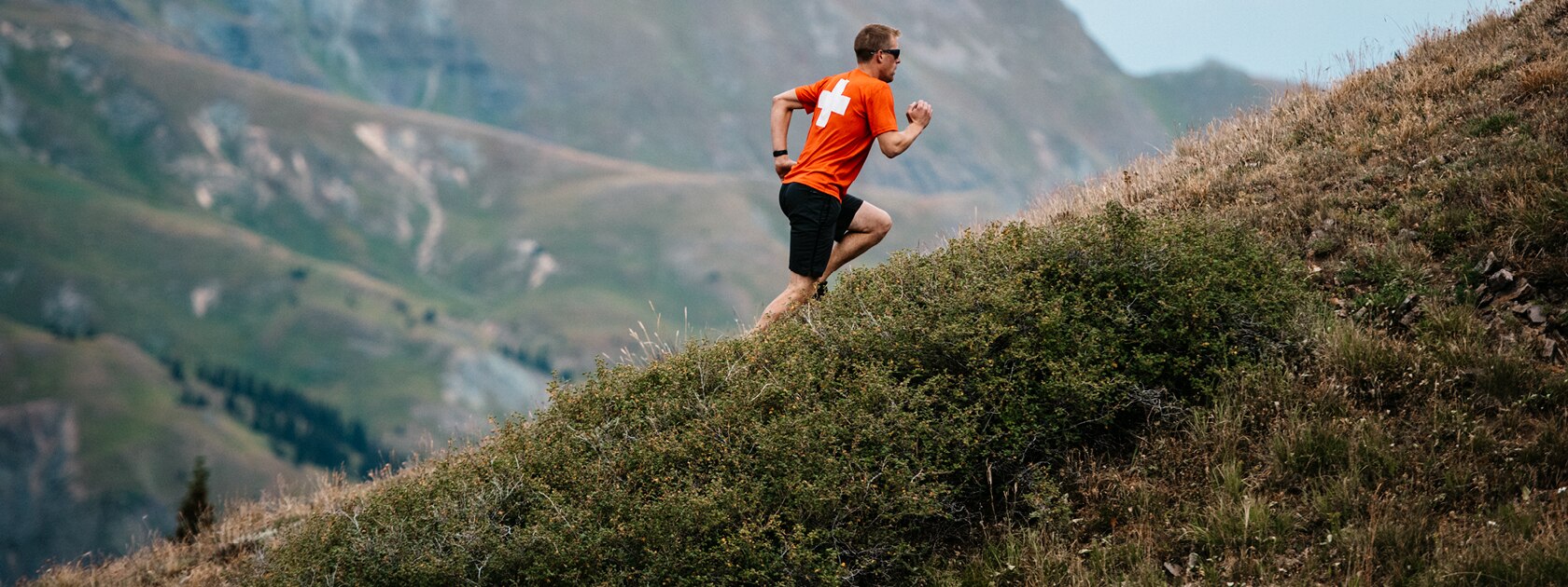 man in orange shirt and black shorts trail runs up a steep grassy slope
