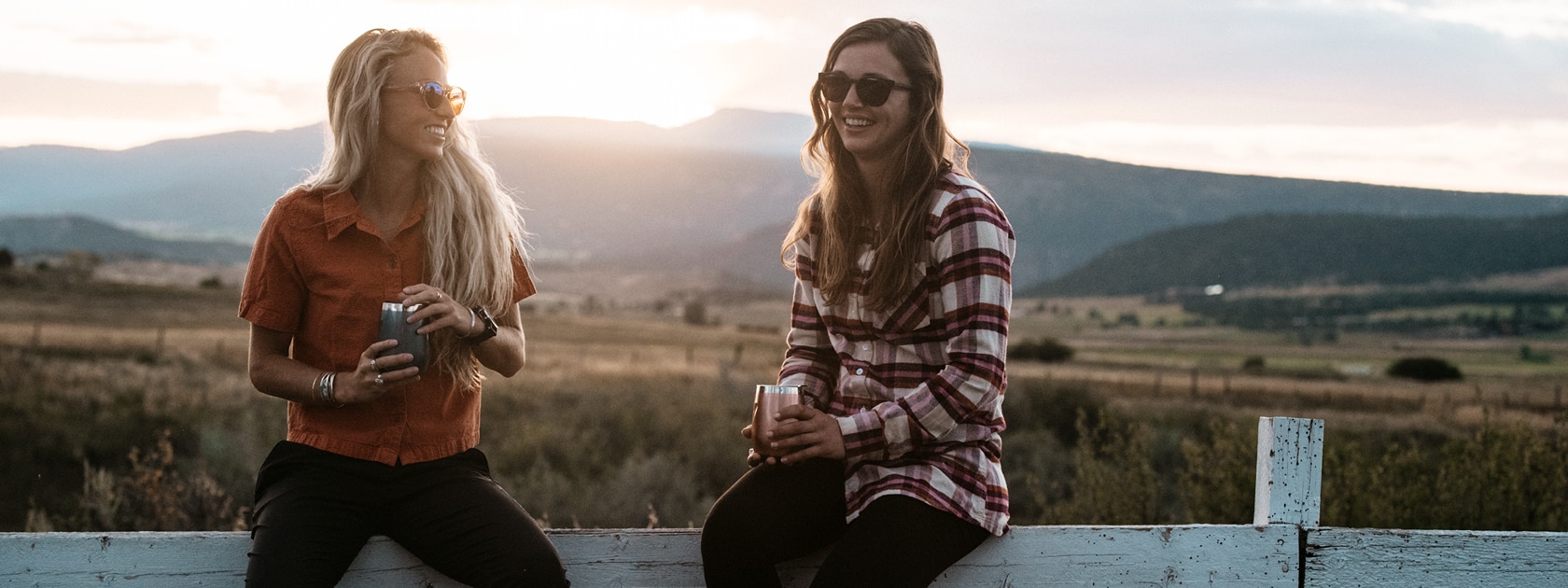 two women holding drinks sit on a white fence while the sun sets over the mountains behind them