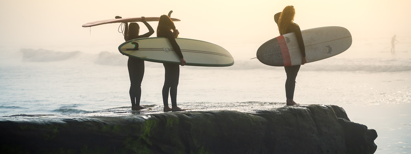 three women holding surf boards look off into the sunny haze of the ocean