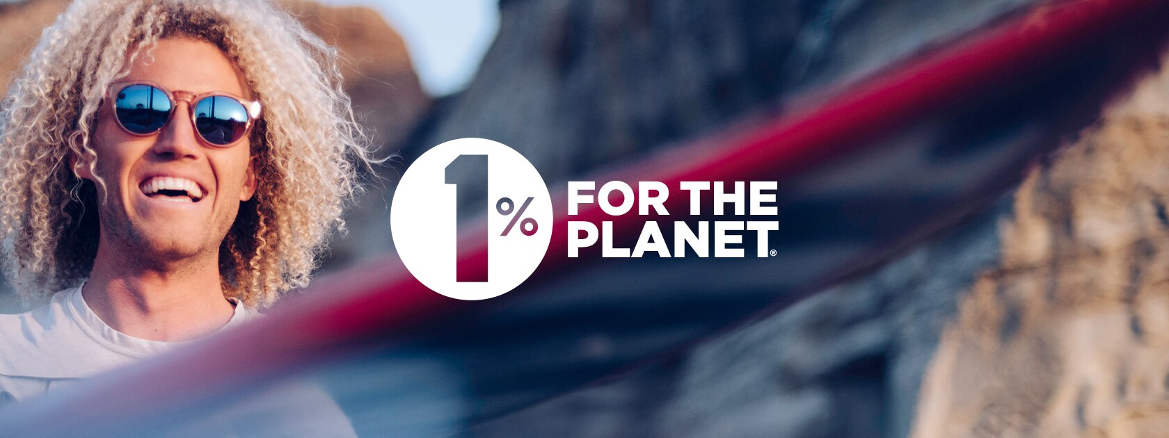 a smiling man wearing sunglasses and holding a surfboard with 1% for the Planet logo in the middle