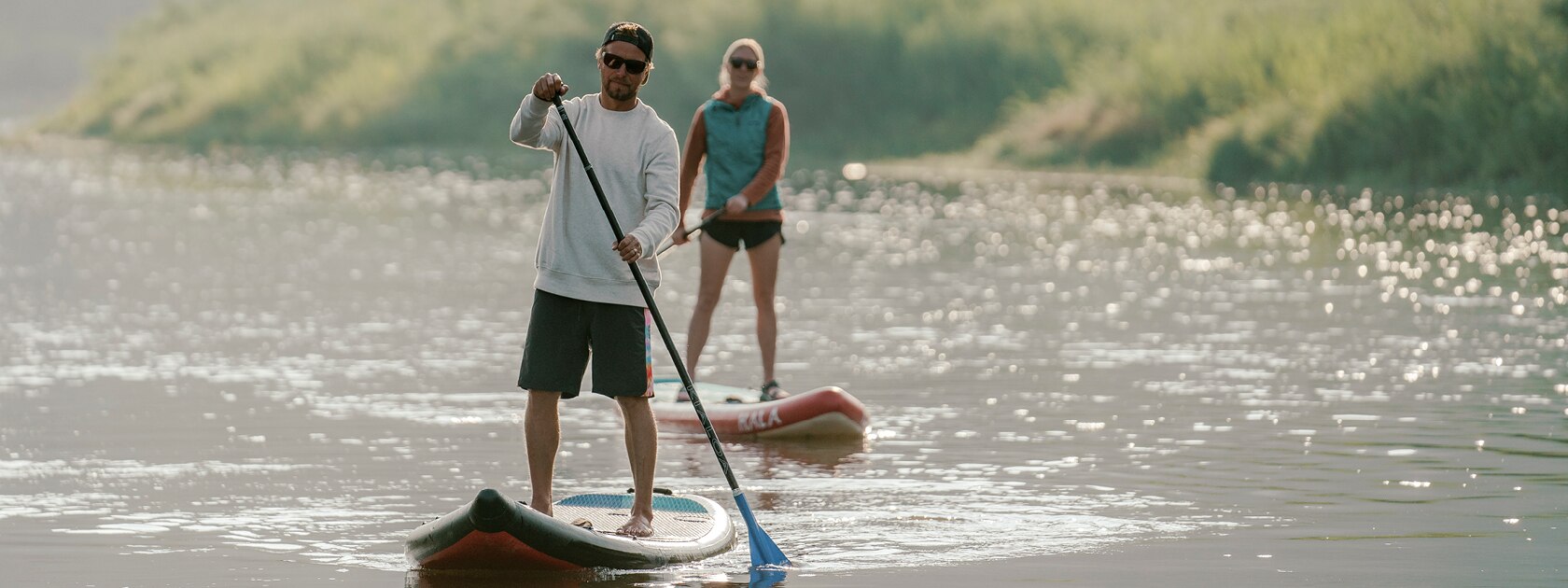 two sunglass wearing stand up paddle-boarders on the water with grassy bank behind them