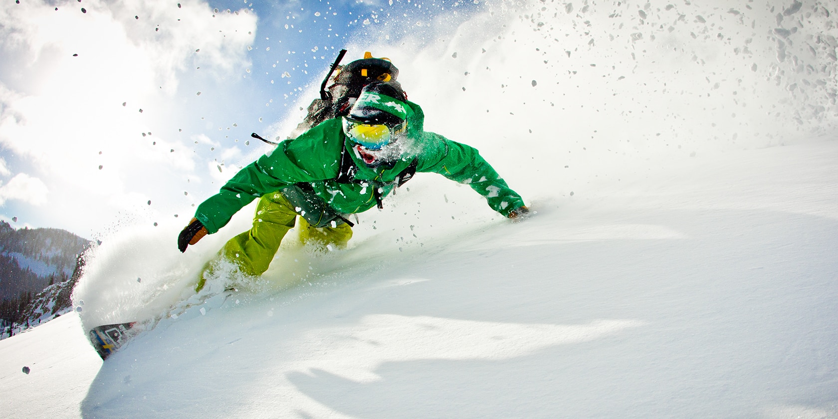 man wearing green snow suit turns fast on a snowboard causing snow to fly into the air