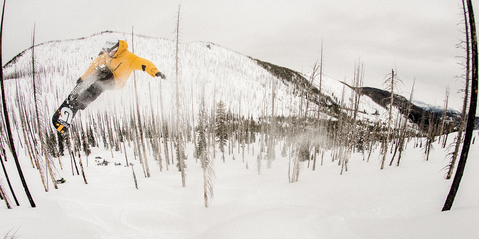 man with yellow coat jumps his snowboard through snow and trees