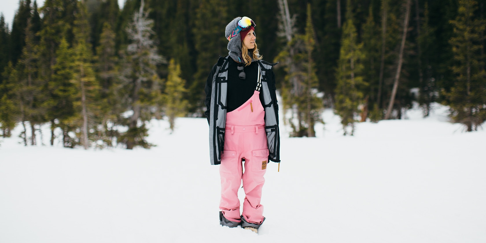 woman with pink snow pants stands in snow with trees in the background