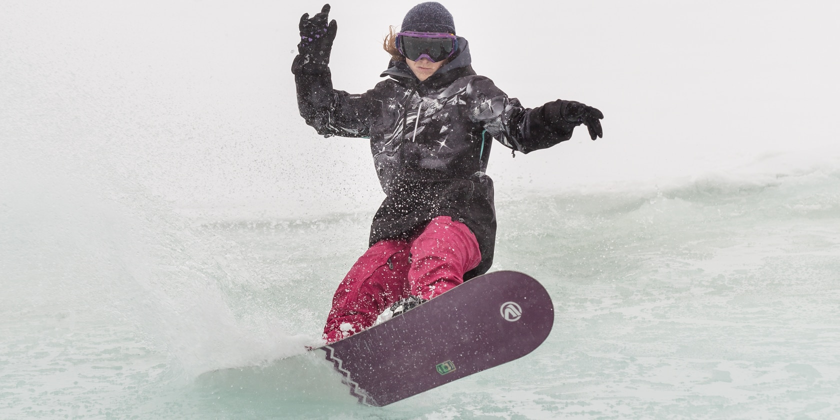 person wearing pink snow pants skims a snowboard over water