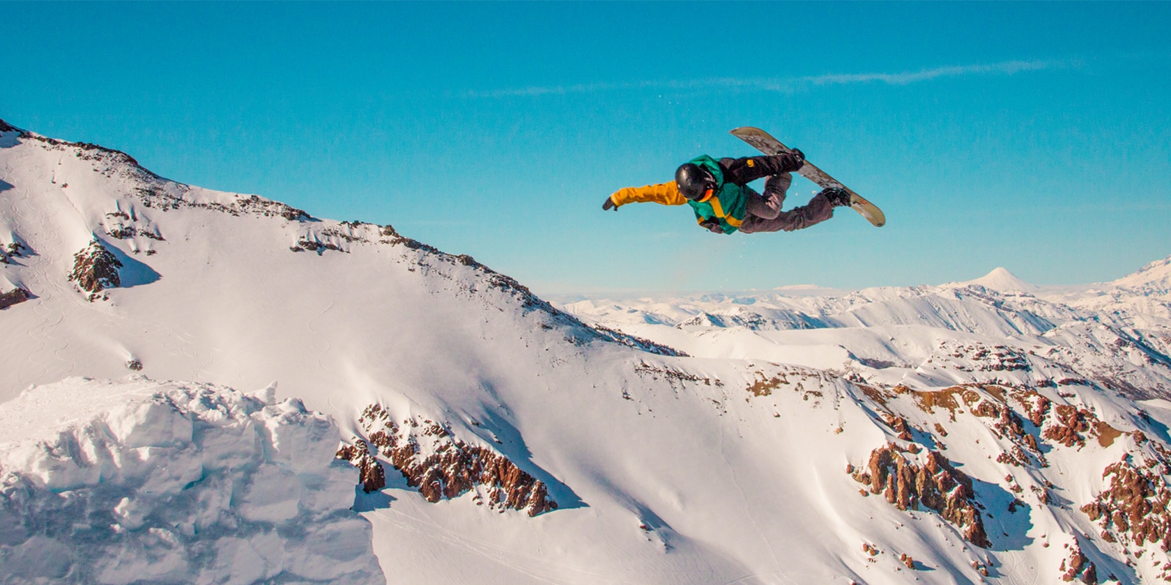 person with green and yellow coat doing flips in the air on a snowboard in front of a mountain background