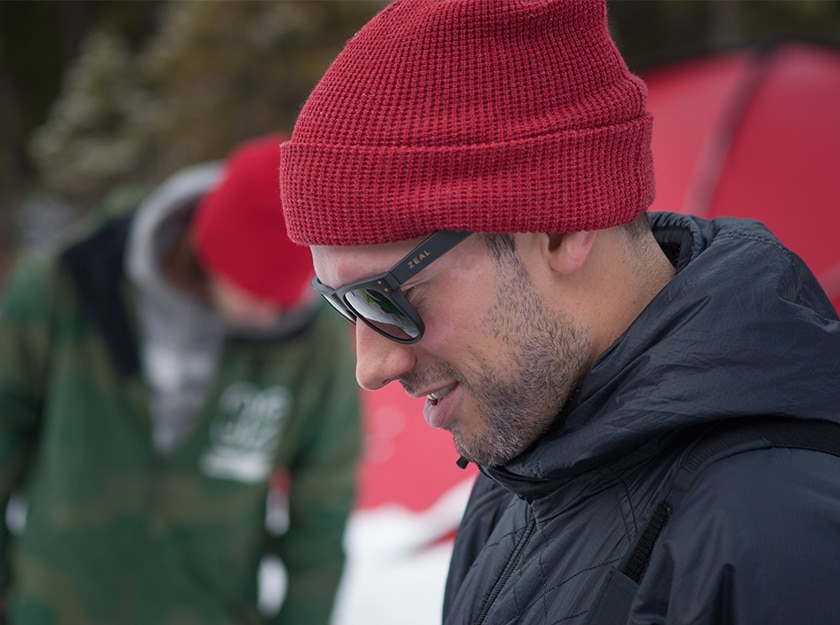 profile of man wearing black coat and red hat and sunglasses with person wearing green coat out of focus in the background
