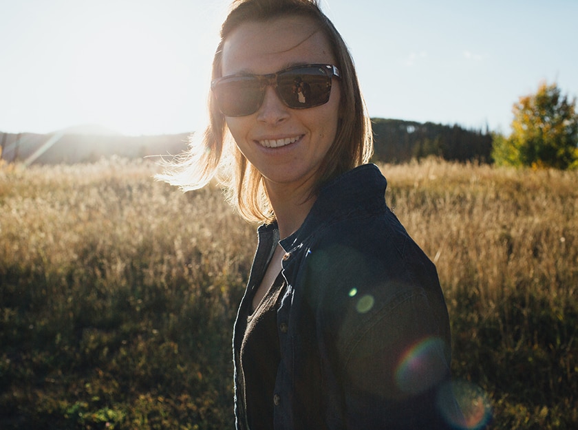 woman wearing black jacket and sunglasses standing in wheat field smiles for camera