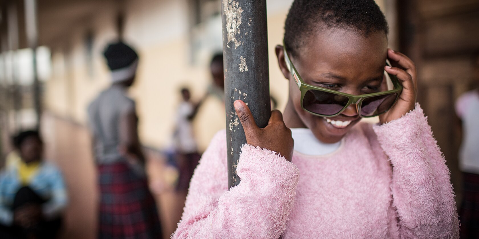young girl wearing pink fuzzy sweater tries on green sunglasses while holding on to post