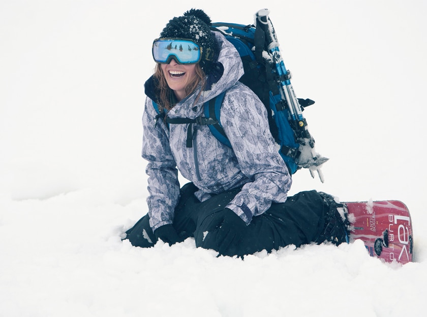 female snowboarder laughing in the snow with goggle with blue lens reflecting pine trees