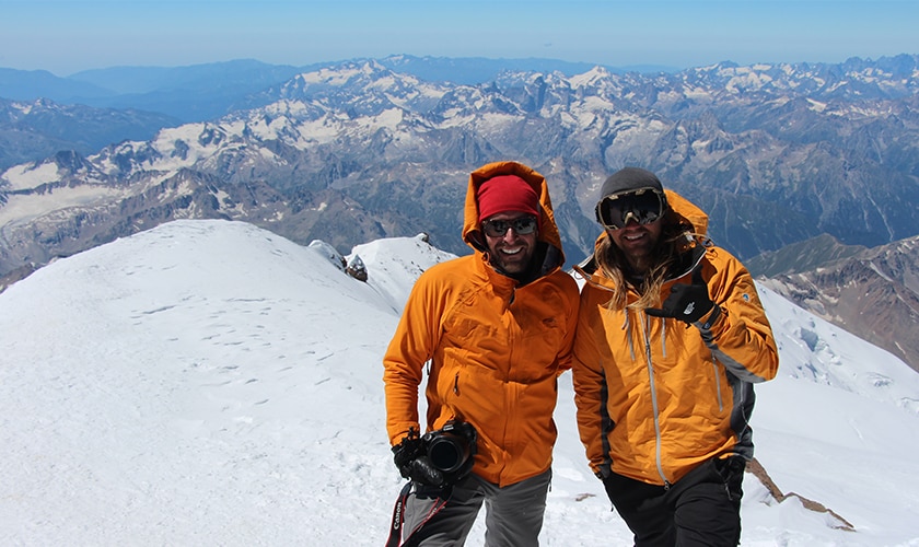 two men with orange coats stand at the top of a snowy mountain peak with mountains in the distance