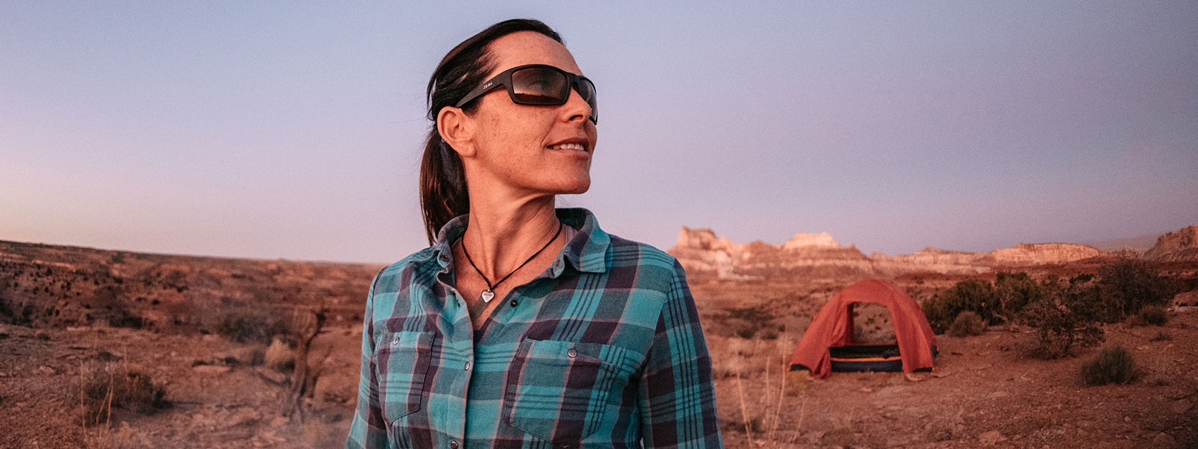 woman in blue shirt and sunglasses standing in the desert near her tent