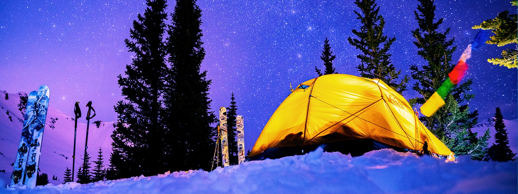a bright yellow tent sits in the snow surrounded by trees and a starry night sky