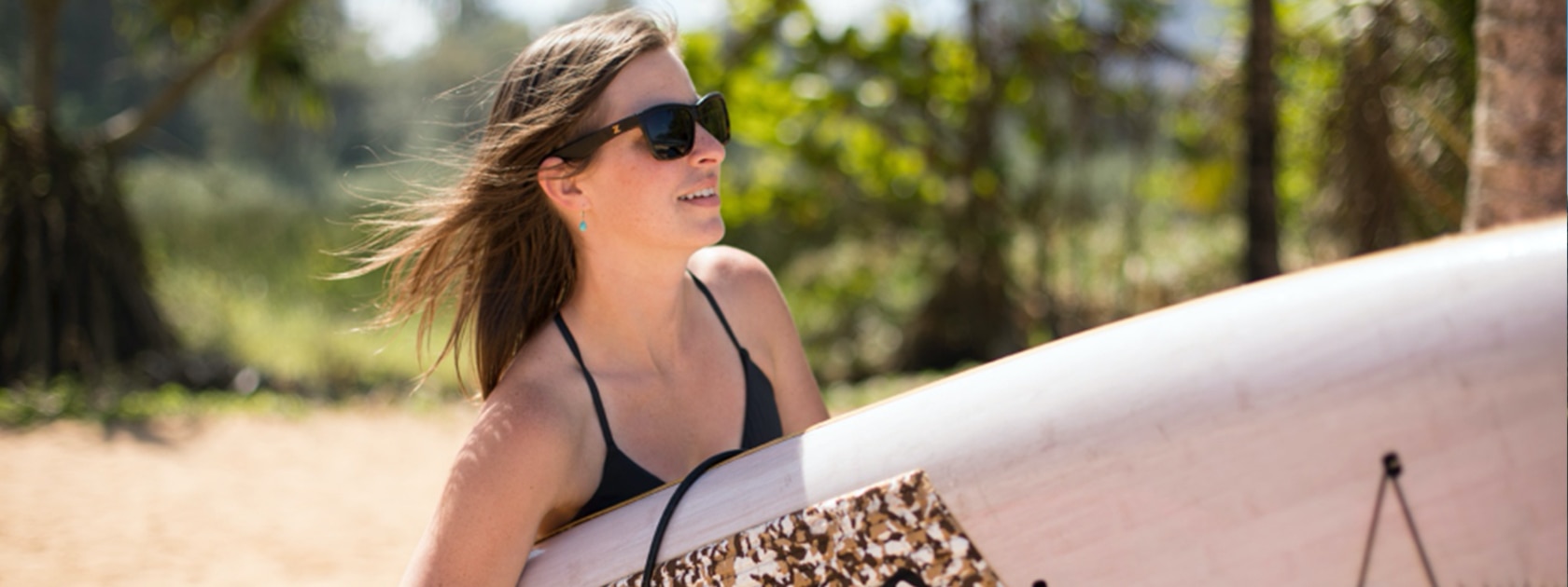 a woman wearing sunglasses and a black top carries a surfboard