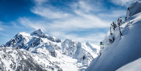 snowboarder making a big jump down a snowy mountain with a mountain and blue sky in the background