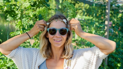 blonde woman with blue frame sunglasses standing in a garden putting a flower crown on her head