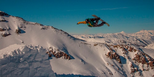 person with green and yellow coat doing flips in the air on a snowboard in front of a mountain background