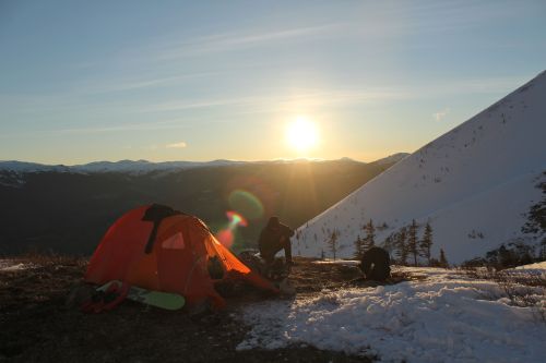 snowboarder sits next to small tent as sun sets over the mountains in the distance