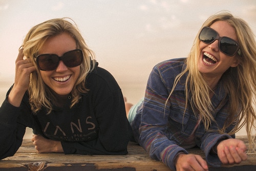 Two blonde women with sunglasses smiling and laughing propped up on elbows