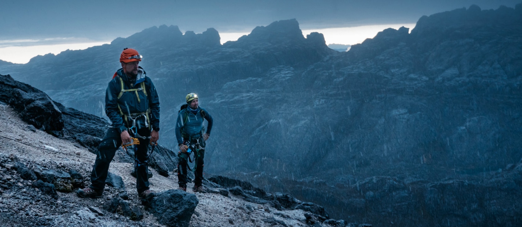 Two men with headlamps and climbing gear stand on a dark mountain as it rains