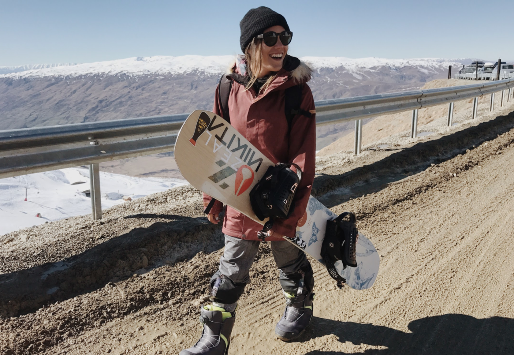 woman wearing red coat and black sunglasses carrying snowboard walks along a dirt road in the mountains