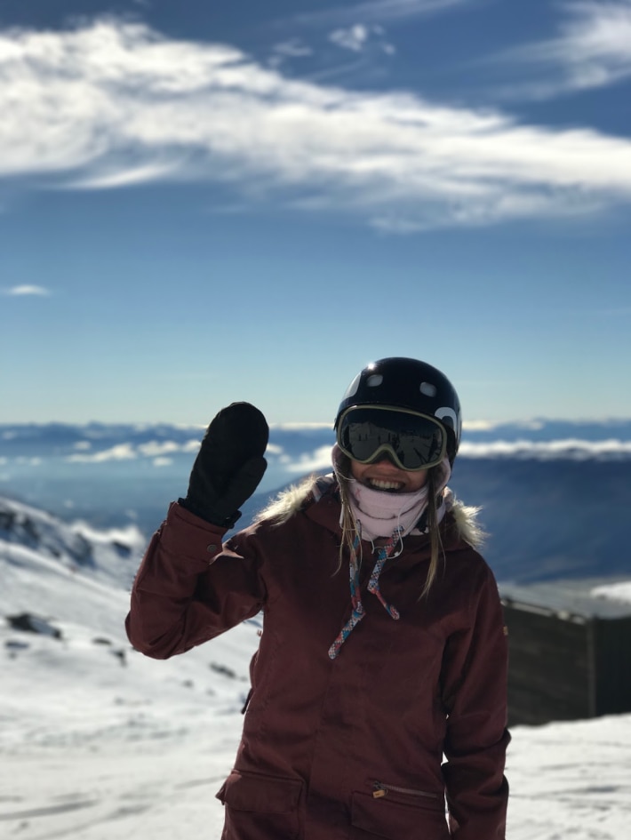 snowboarder standing in snow wearing green frame black lens goggles smiling and waving with mountains in the background