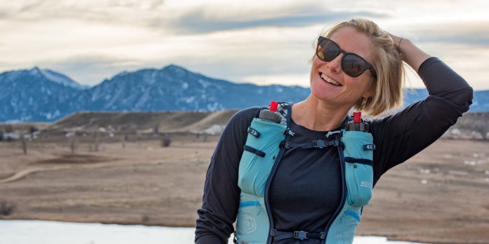 Blonde smiling woman with her hand in her hair wearing blue hiking backpack and sunglasses