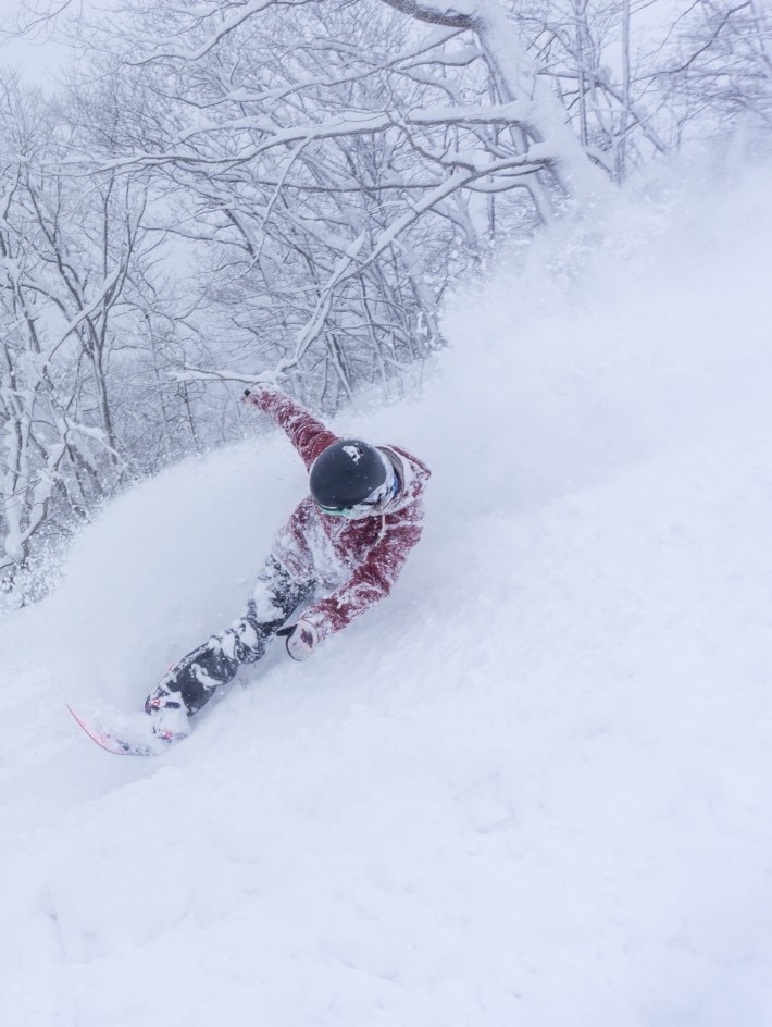 snowboarder in red coat leaning wildly and spraying snow as she rides down mountain