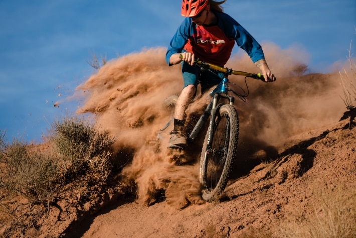 Mountain biker in red helmet kicks up red dust as he turns on a dirt hill