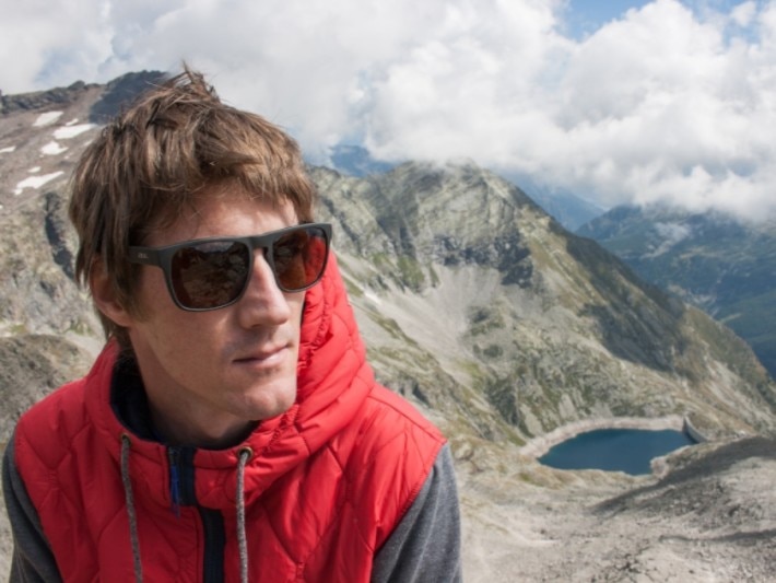 close up of man in red coat and black sunglasses with a lake and mountains in background 