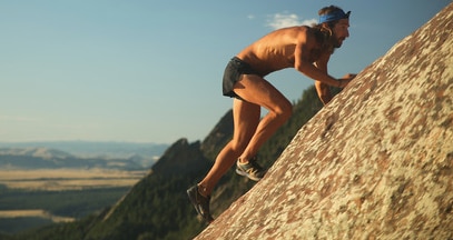 Man with long brown hair and beard climbing up a flat mountain surface in the sun