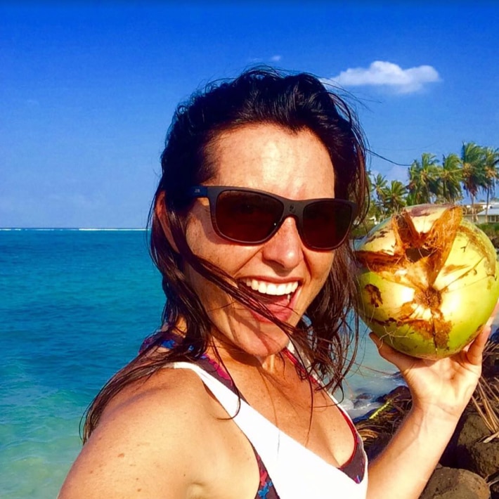 Woman wearing sunglasses smiling on a beach holding a coconut