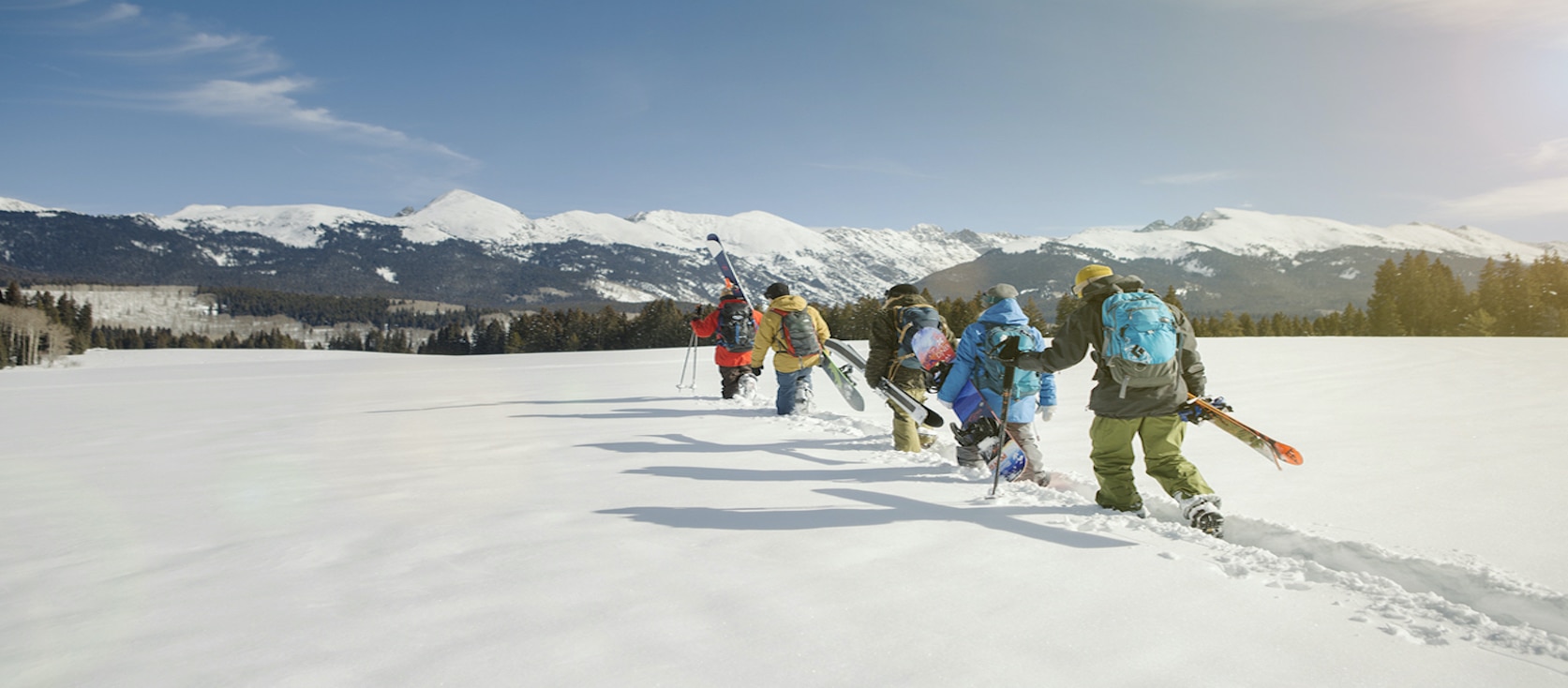 five skiers holding their skis walking through the snow with mountains in the background
