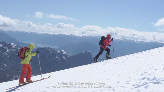 two skiers walking up a slope with mountains in the background
