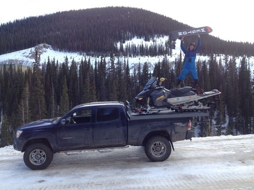 man holding a snowboard over his head while standing on a snow mobile in the trunk of a truck parked in the mountains