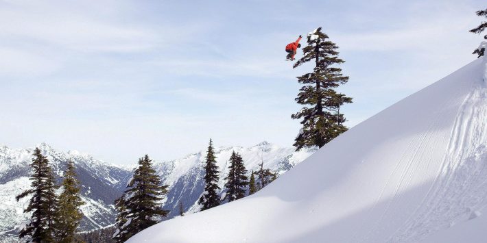 snowboarder flying through the air over snowy mountain with trees behind