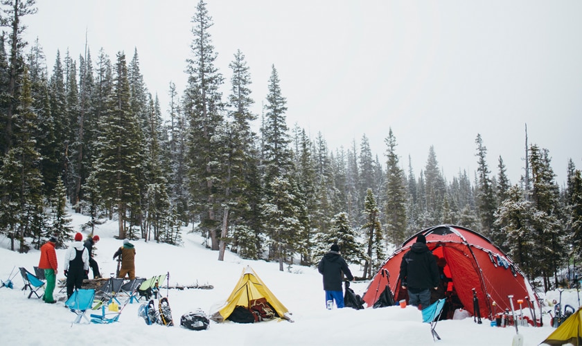 group of people campers pitching tents in the snow at the edge of a forest