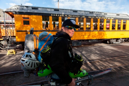 Skier in black cap and sweatshirt carrying all his gear in front of a yellow train car
