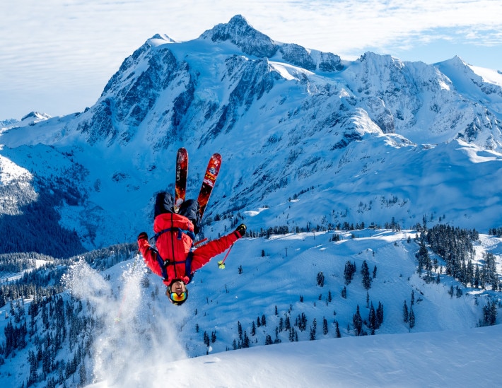 skier in red coat flipping off a jump with a mountain in the background