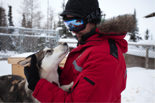 man in red coat and blue lens ski goggles petting a husky dog in the snow