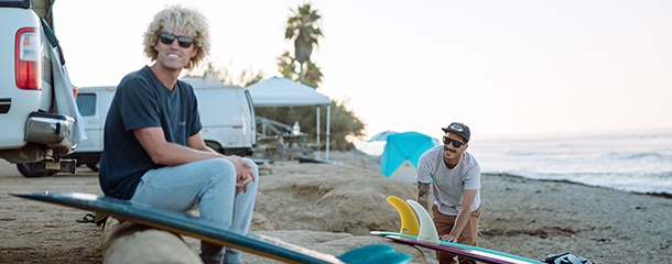 two men sitting on the beach with their surf boards wearing sunglasses