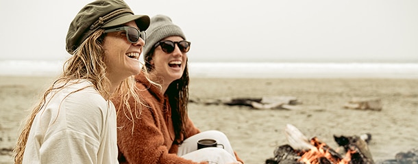 two women laughing and sitting on the beach wearing hats and sunglasses